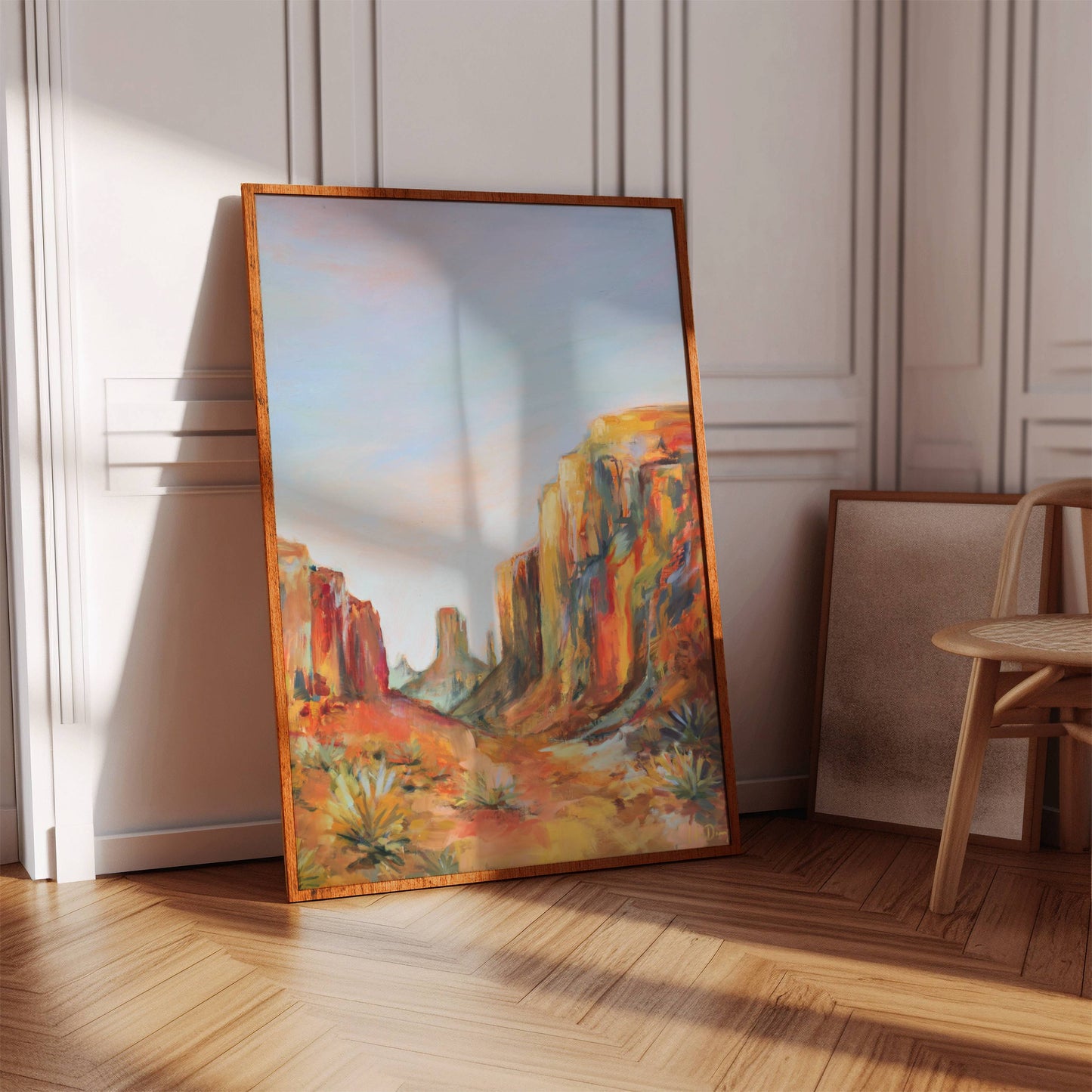 Framed painting of an Arizona desert landscape on a wooden floor with a chair in the background.