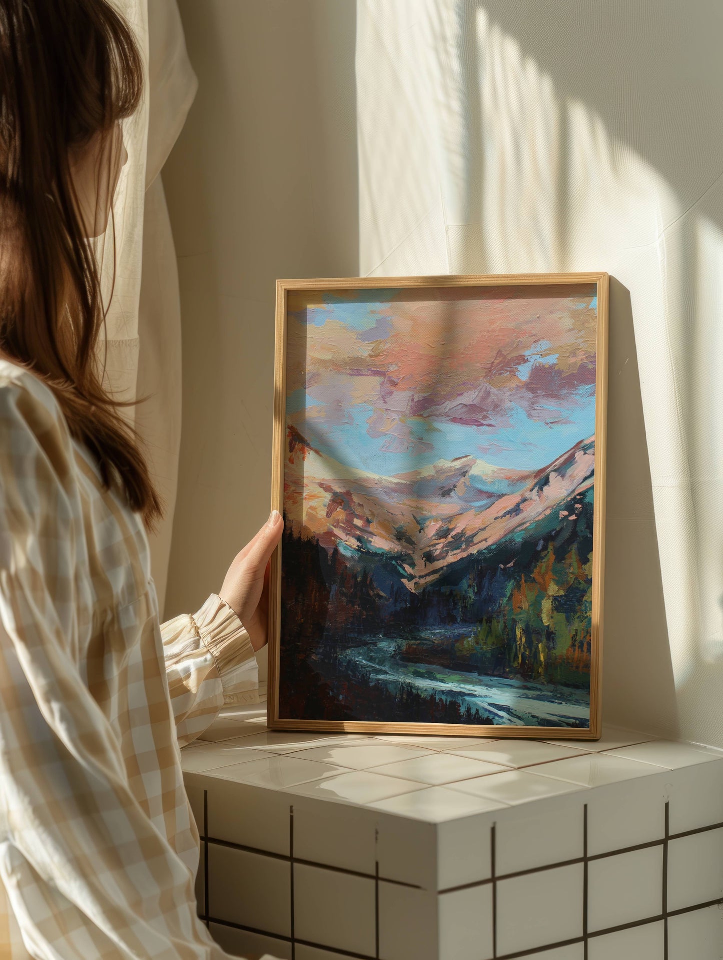 Person holding a framed painting of a mountain landscape in a softly lit room.