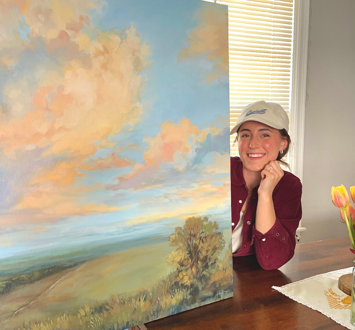 Woman standing next to a large landscape painting on a wall.