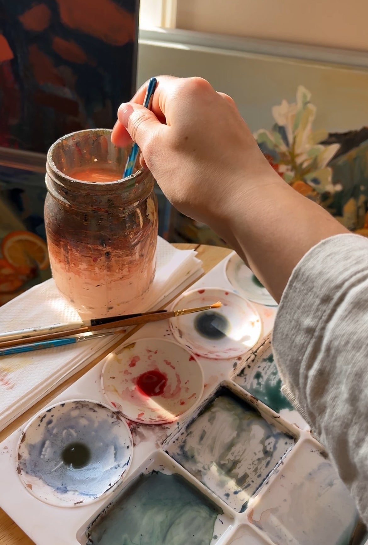 Person painting with a brush in a jar of paint next to a palette with various colors.