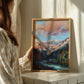 Person holding a framed painting of a mountain landscape in a softly lit room.