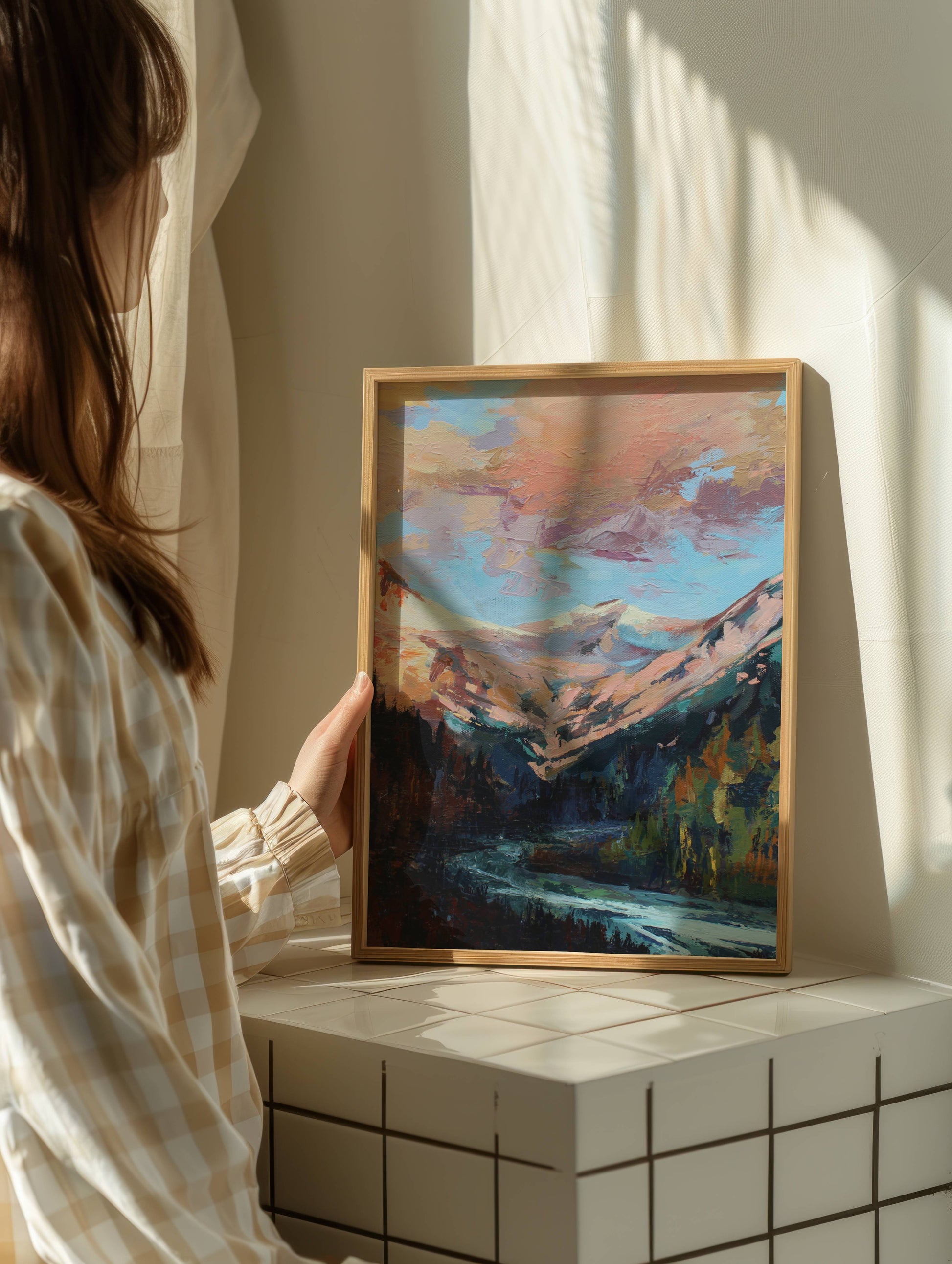 Person holding a framed painting of a mountain landscape in a softly lit room.