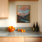 Kitchen counter with a bowl of oranges, scattered oranges, and wine bottles, with a mountain painting on the wall.